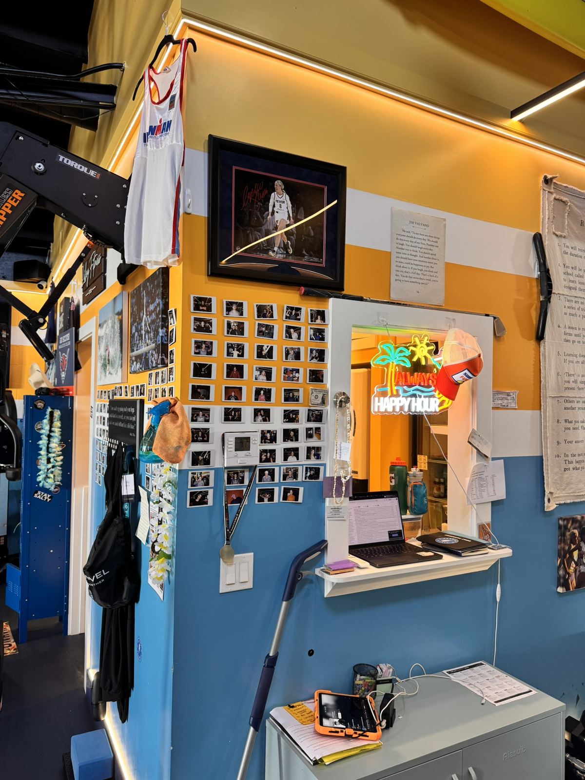 The front desk corner at Lilley Fitness, with a wall of member photos and an Always Happy Hour neon sign.