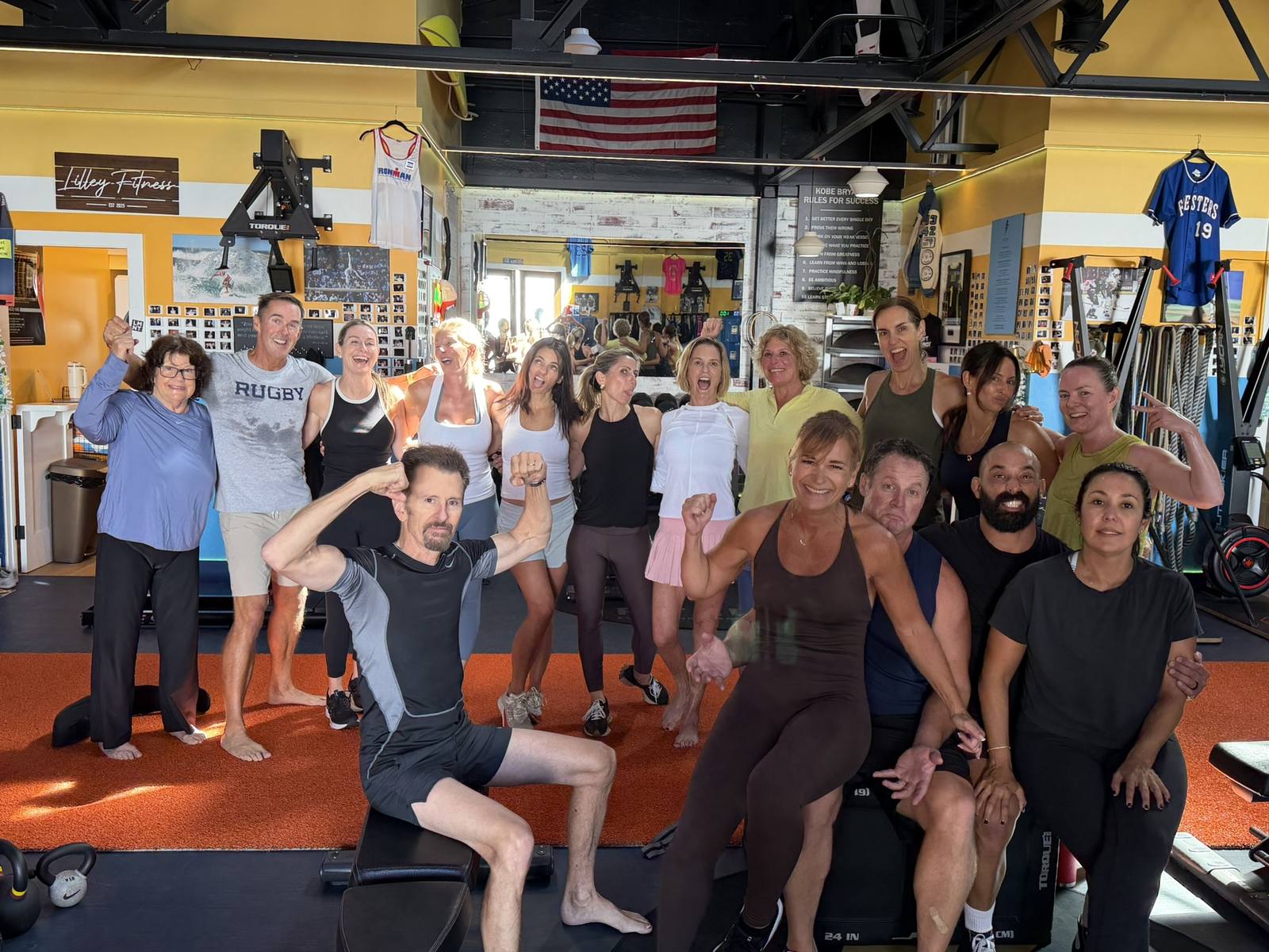 A big group of Lilley Fitness members of all ages flexing and smiling under the Lilley Fitness sign.