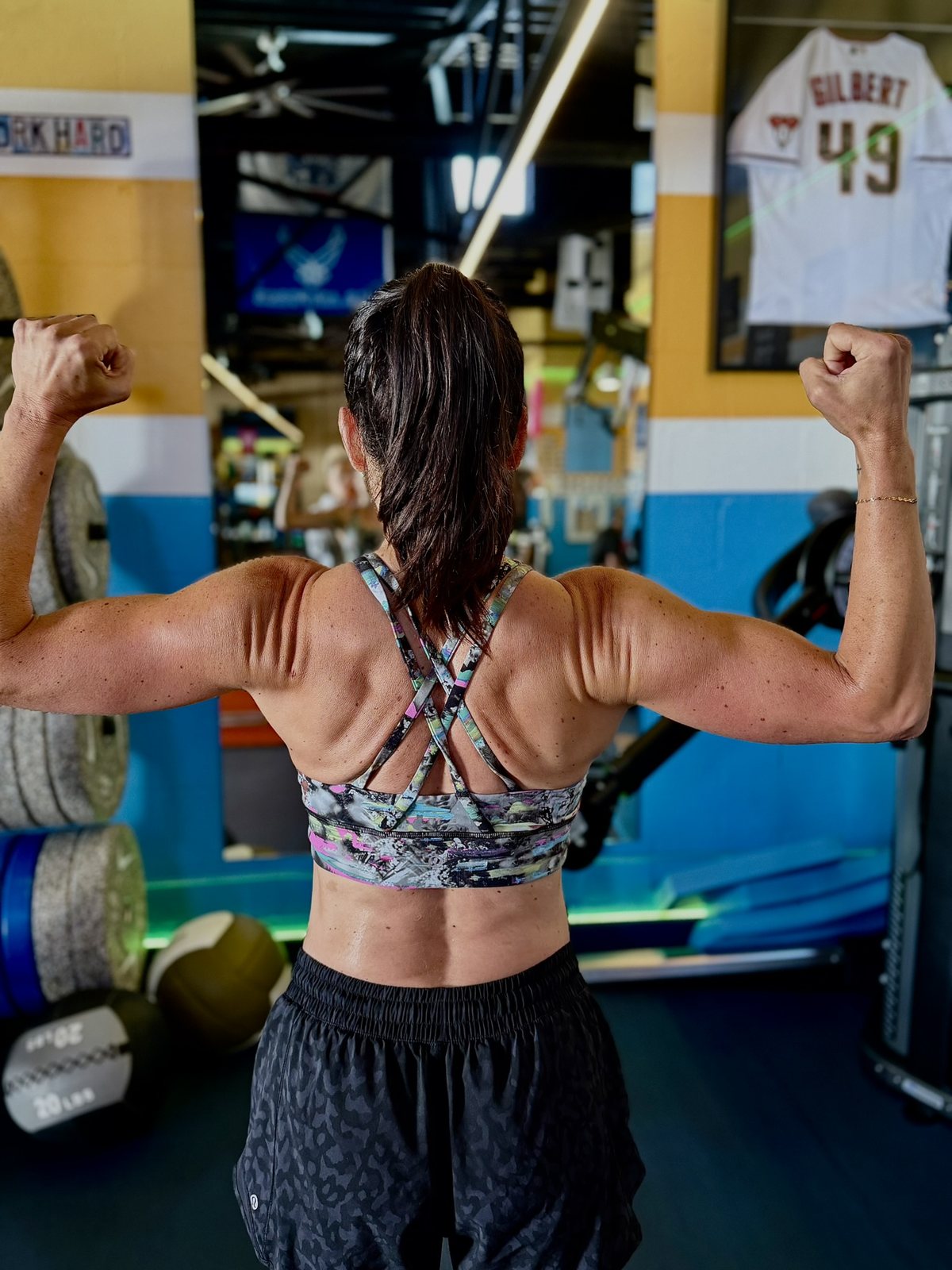 A member seen from behind, flexing her back and arms beside the weights and a framed jersey.